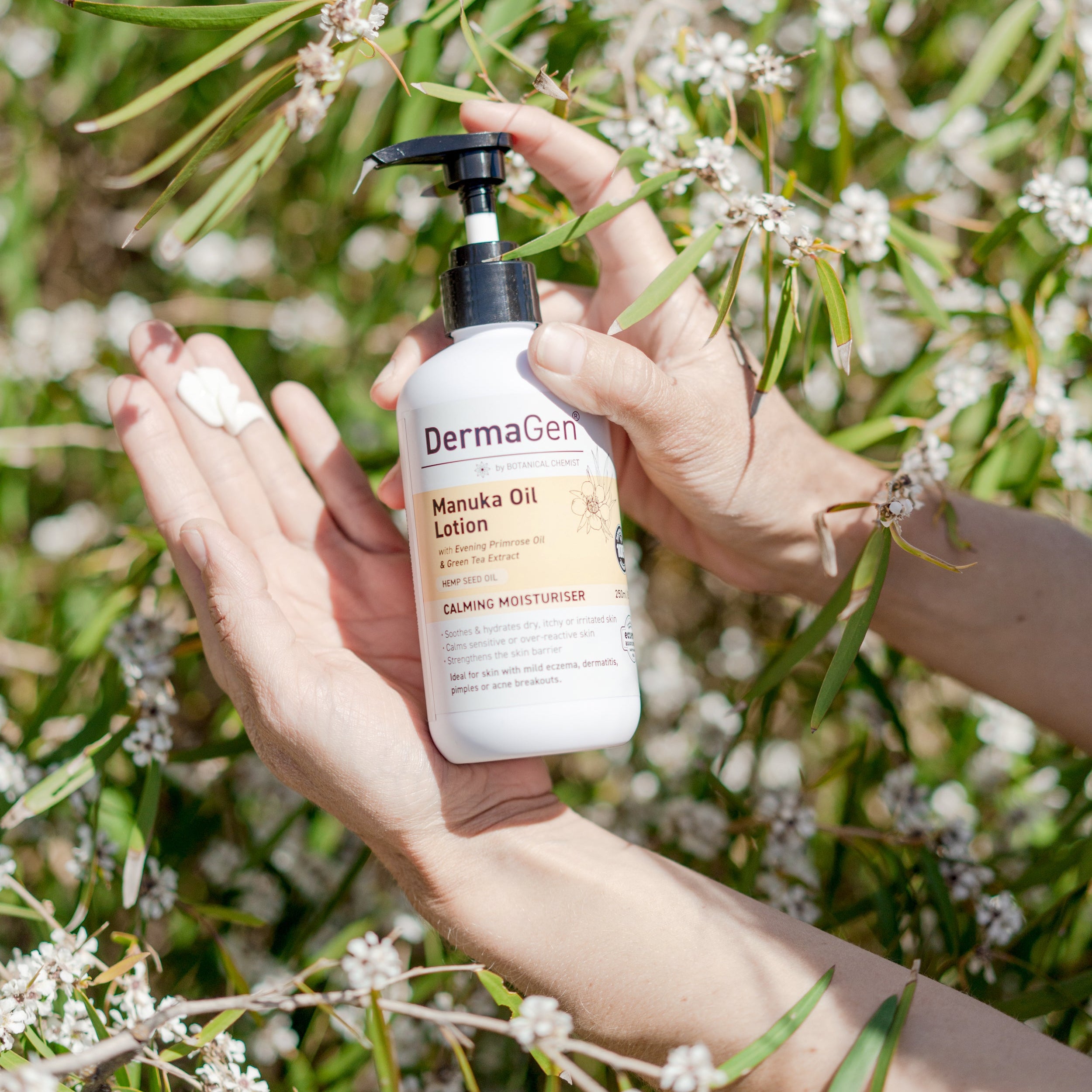 Person holding a bottle of DermaGen Manuka Oil Lotion in front of green foliage.