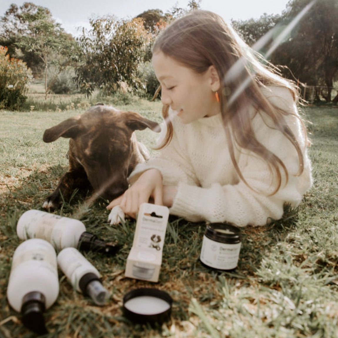 Child sitting on grass with a dog, surrounded by skincare products
