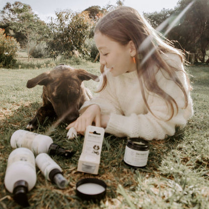 Child sitting on grass with a dog, surrounded by skincare products