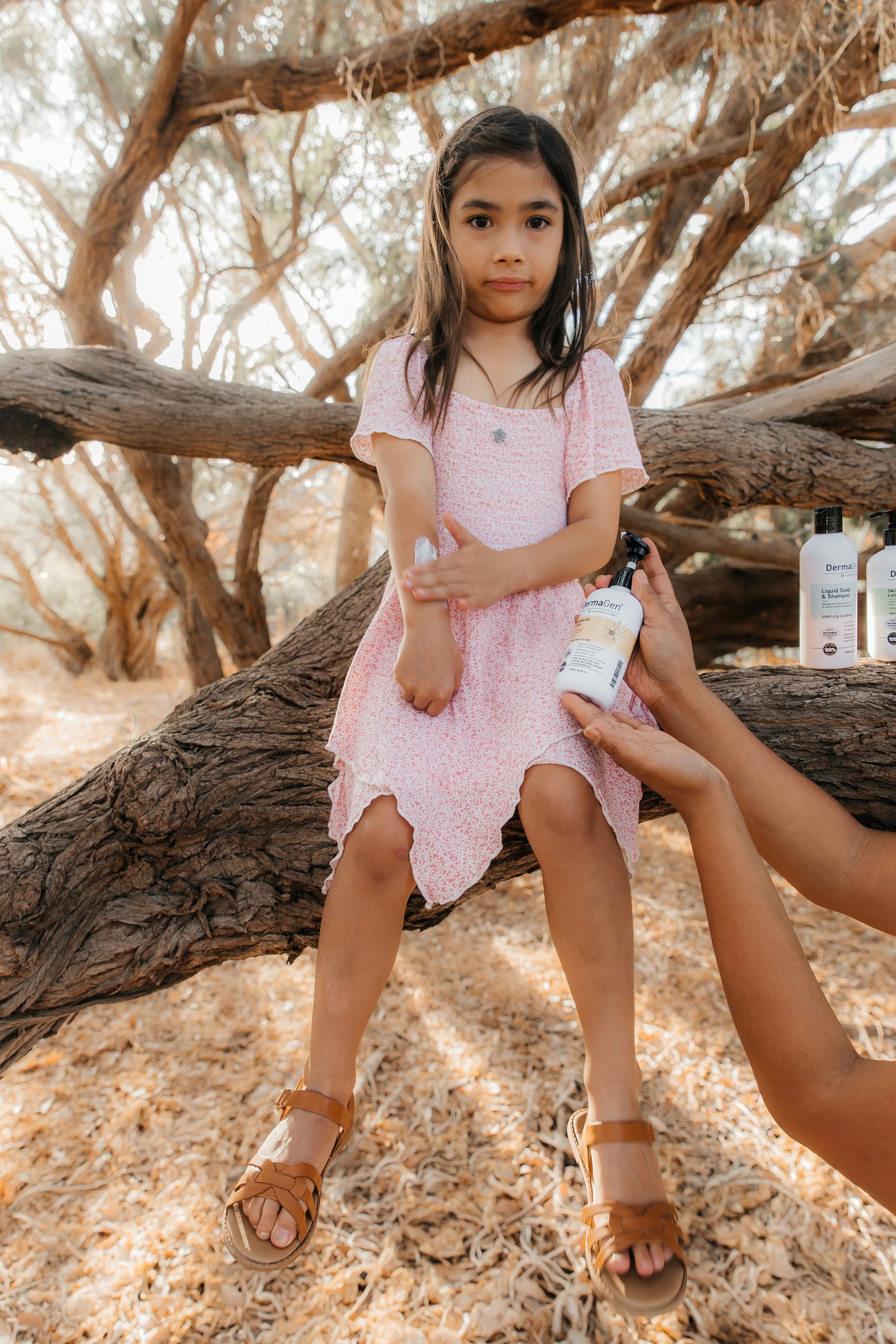 Young girl in a pink dress sitting on a log outdoors, with a bottle of lotion held by an adult.